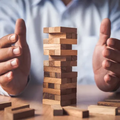 Hände um einen Turm aus Holzklötzen auf einem Tisch, als Symbol für Stabilität, Balance oder Risiko.
