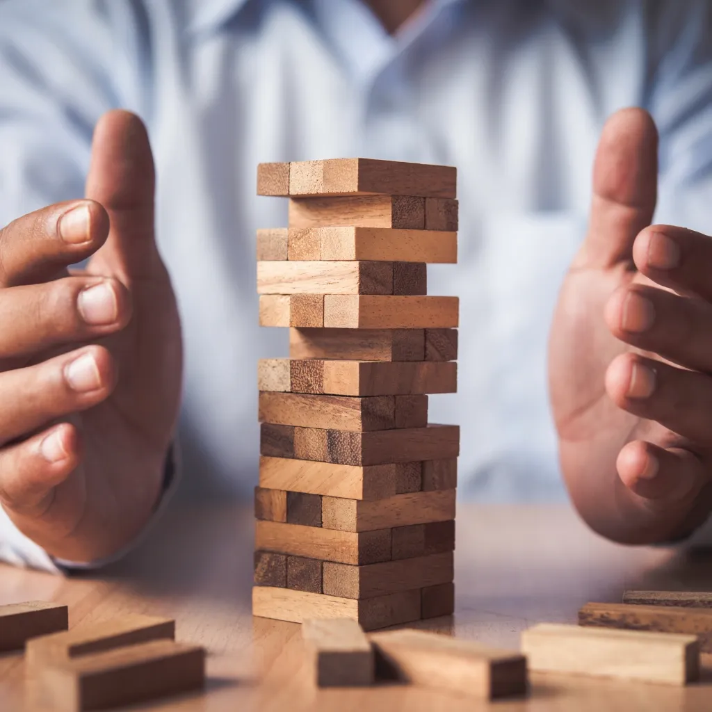 Hände um einen Turm aus Holzklötzen auf einem Tisch, als Symbol für Stabilität, Balance oder Risiko.