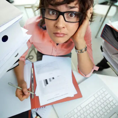 Stressed office worker sitting between tall stacks of folders at a desk, holding a pen and looking overwhelmed into the camera, with documents and a laptop in front of her.