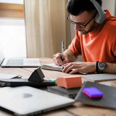 A man with headphones writes in a notebook at a desk.