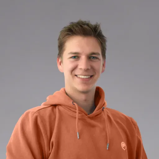 Ein junger Mann mit blauen Augen lächelt in einem orangen Hoodie.