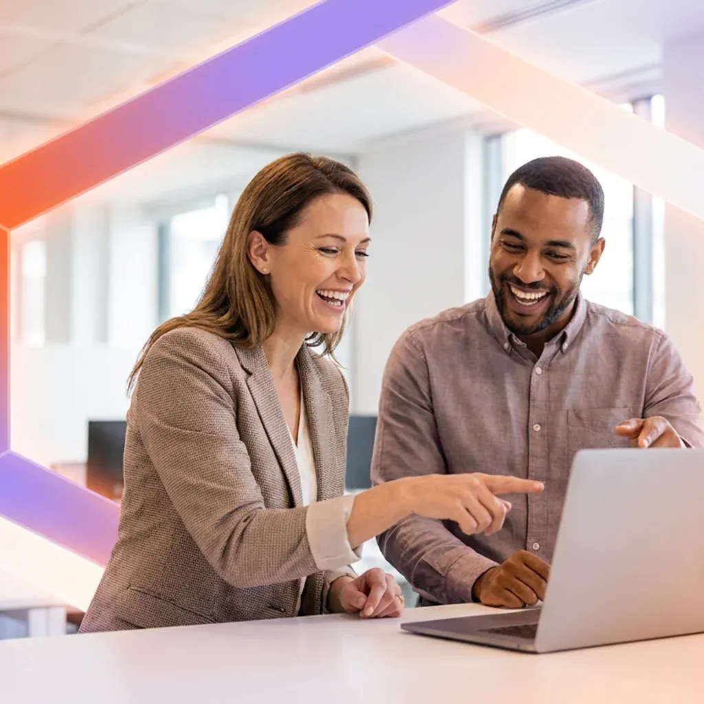 Two smiling colleagues pointing at a laptop screen in a modern office.