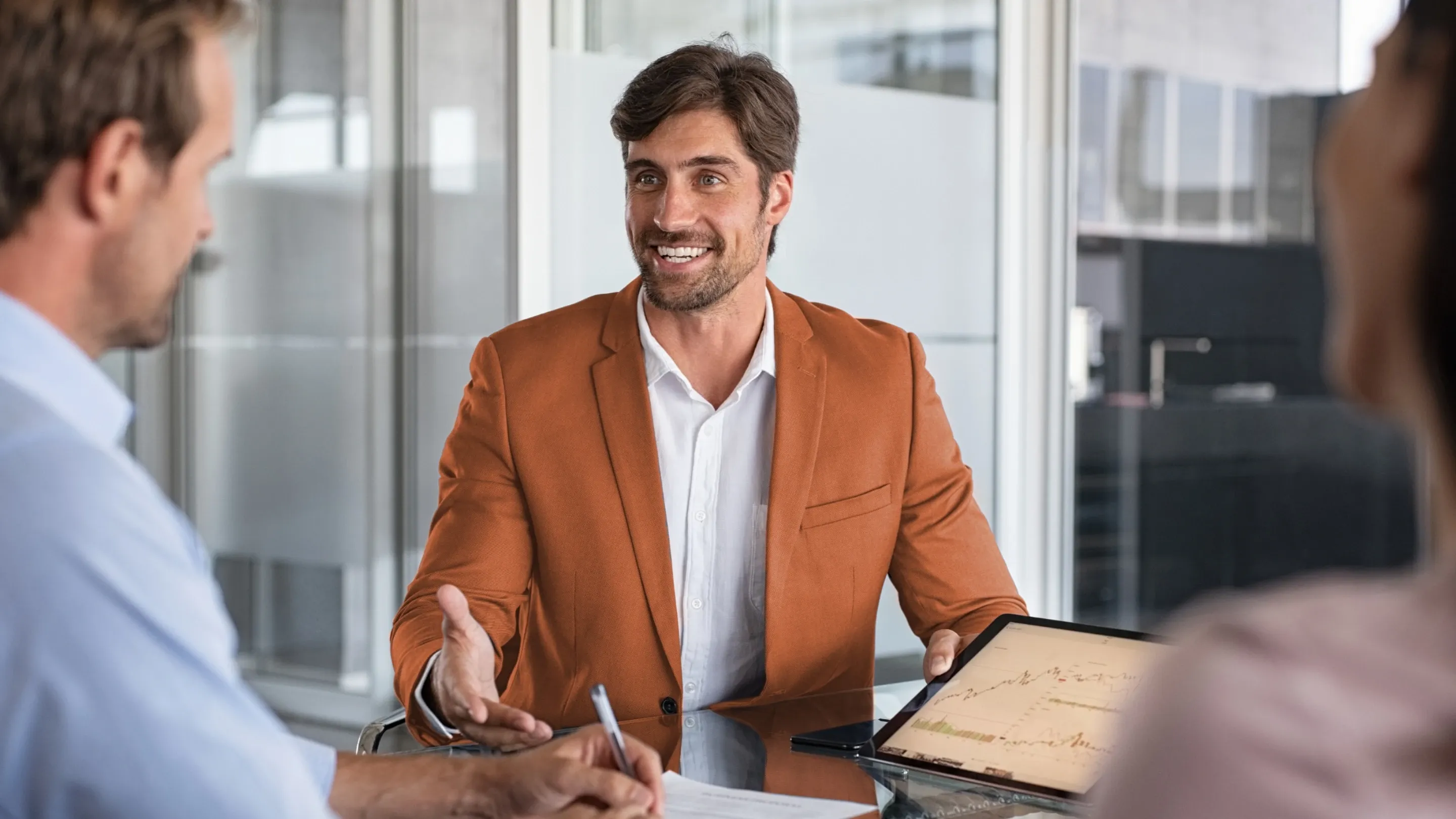 A smiling man in a blazer presents a chart on a tablet.