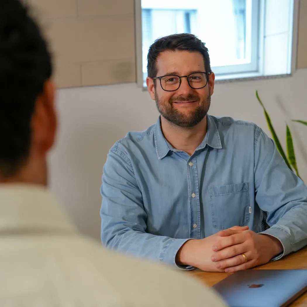 Ein Mitarbeiter sitzt an einem Schreibtisch und lächelt einen Kollegen während einer Besprechung an.