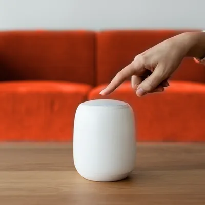 Ein Finger berührt einen weißen Smart Speaker auf einem Tisch.