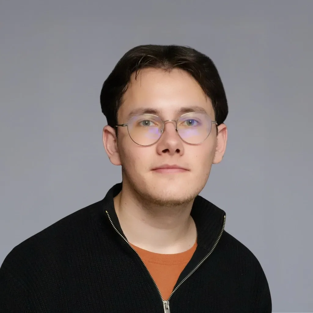 Junger Mann mit Brille in einer schwarzen Reißverschluss-Strickjacke.