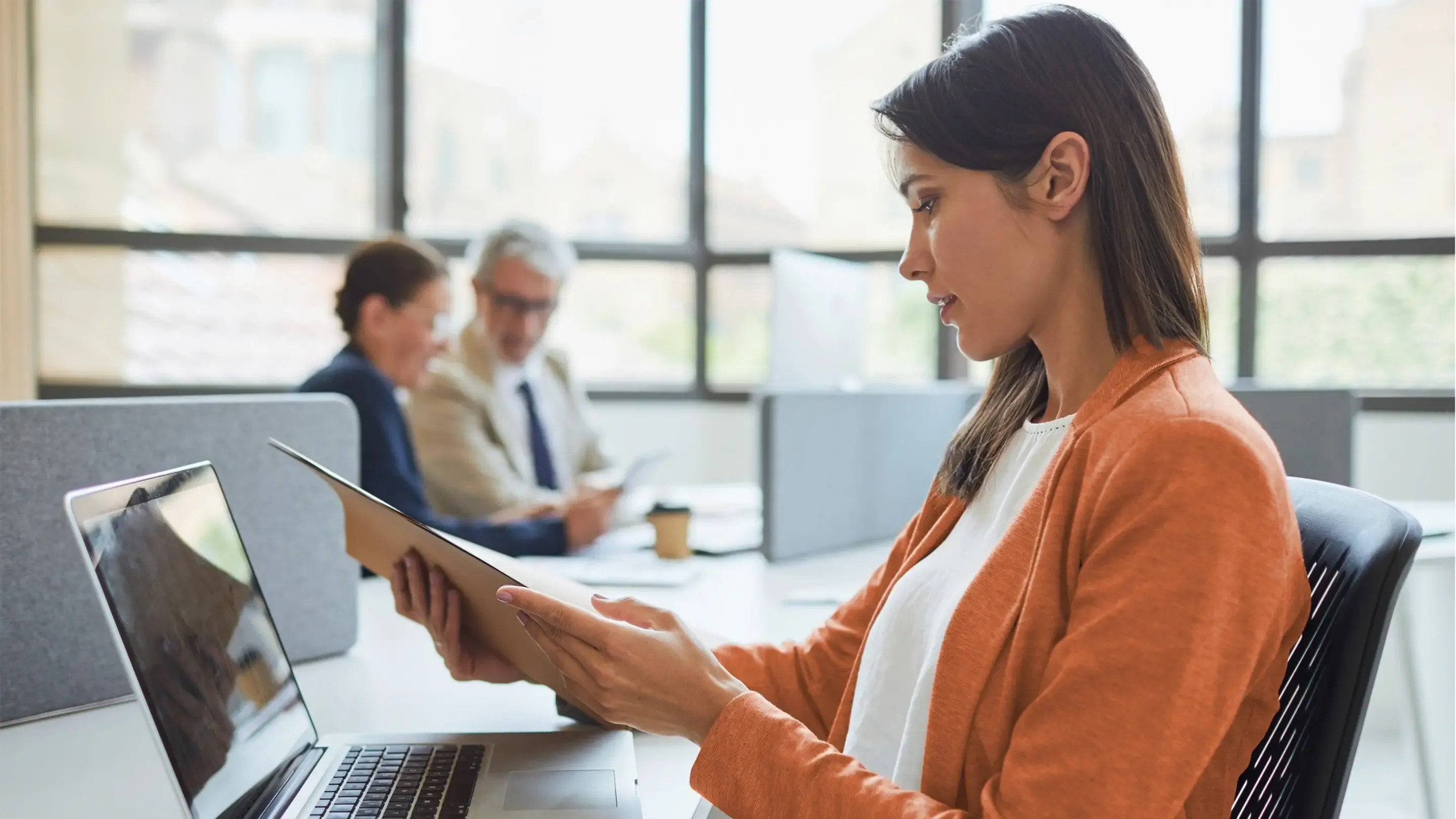 A woman in an orange cardigan reviews a folder at her desk.