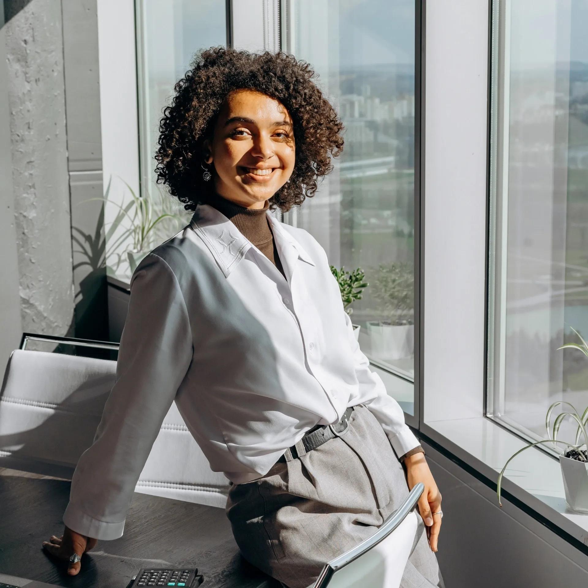 A smiling person with curly hair leans against a desk.