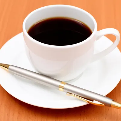 Cup of black coffee on saucer with ballpoint pen next to it.